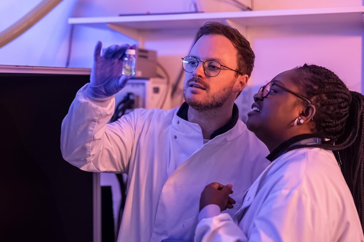 Two researchers wearing lab coats are closely observing a vial.